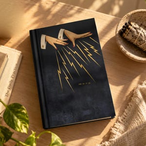 May include: A black hardback journal featuring a graphic illustration of two hands with red nail polish extending from white cuffs, emitting golden lightning bolts downwards. The text AND SO IT IS is printed in small gold letters below the lightning.