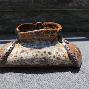 May include: A handcrafted ceramic vase with a unique design. The vase features a brown rim and accents, with a speckled beige and brown body. Decorative brown side panels with cut-out details add to its artistic appeal.