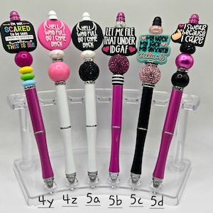 May include: Seven pink, black, and white pens with colorful beads and charms. The pens have different sayings on the charms, including "I'm not scared to be seen", "Hell, who full, go I came back", "Let me file that under IDGAF", "My neck, my back, my anxiety attack", and "I swear because I care".