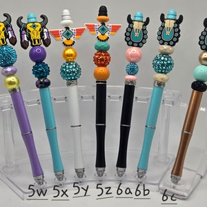 May include: Seven colorful pens with different decorative tops, including a turquoise skull, a yellow skull, a turquoise and orange bird, a turquoise and orange bird, a turquoise skull, a turquoise skull, and a turquoise skull. The pens are arranged in a clear plastic holder.