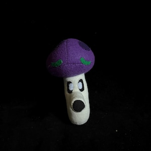 May include: A plush toy mushroom with a purple cap, green accents, and a white stem. The mushroom has large, cartoonish eyes and a black mouth. The toy is made of soft fabric and is designed to resemble a character from a video game.