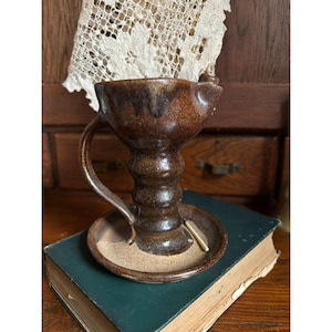 May include: A handmade brown ceramic candle holder with a handle and a small dish. The candle holder has a textured surface and a unique shape. The candle holder is sitting on a green book.