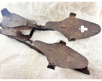 May include: A pair of antique ice skates, constructed from aged metal. The skates exhibit a dark brown, rusty finish, with a clover-shaped cutout on each blade. The blades are linked by a metal bar, designed to be affixed to footwear.