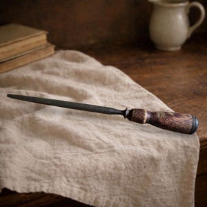 Puede incluir: Una chaira de metal oscuro con un mango de madera. El mango tiene un patrón de vetas de madera marrón oscuro. La chaira está sobre un paño de lino beige, con un fondo borroso de una mesa de madera y una jarra de cerámica.