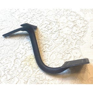 Könnte beinhalten: Ein schwarzes Metallfußpedal mit einem gebogenen Arm und einer flachen, rechteckigen Trittfläche. Das Pedal ist für die Verwendung mit einer Maschine oder einem Gerät konzipiert.