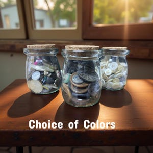 May include: Three glass jars filled with a variety of colorful buttons. The jars have cork lids and are sitting on a wooden surface. The text "Choice of Colors" is visible below the jars.
