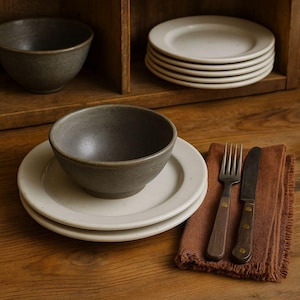 Puede incluir: Un juego de comedor rústico con un cuenco de cerámica gris sobre un plato blanco, con un tenedor y un cuchillo sobre una servilleta marrón. Platos y un cuenco adicionales están apilados en un estante de madera. La vajilla está sobre una superficie de madera.
