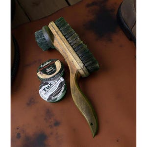 Könnte beinhalten: Eine Holzschuhbürste mit grünen Borsten und einem gebogenen Griff, zusammen mit zwei Schuhcremedosen. Eine Dose ist mit "Griffin Basic Black" und die andere mit "Tux Renovating Polish Dark Brown" beschriftet. Die Artikel befinden sich auf einer braunen Lederoberfläche.