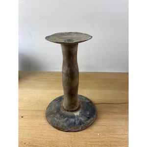 May include: A vintage metal candle holder with a wooden base. The holder has a circular base and a tall, cylindrical stem. The surface is weathered and shows signs of age.