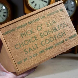 Könnte beinhalten: Eine hellbraune Holzkiste mit dem grünen Aufdruck "PICK O' SEA CHOICE BONELESS SALT CODFISH". Die Kiste hat eine leicht raue Textur und einen Vintage-Look. Der Text ist in einer klassischen Schriftart gehalten.