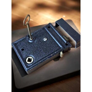May include: A black antique lock with a key inserted. The lock is rectangular with a textured surface and a keyhole. The key is made of metal and has a circular handle. The lock is on a dark brown surface.