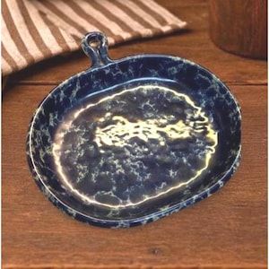 Può includere: Un piatto rotondo in ceramica con una finitura smaltata marmorizzata blu scuro e bianca. Il piatto presenta una piccola impugnatura circolare sul bordo superiore e una superficie interna leggermente strutturata e irregolare che crea profondità.