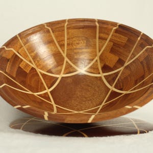 May include: A wooden bowl with a geometric pattern of light and dark wood. The bowl is sitting on a wooden base.