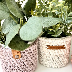 May include: Two potted plants with crocheted covers. The plant on the left has a pink cover with a brown leather tag that says "Loved". The plant on the right has a white cover with a brown leather tag that says "You are so loved".