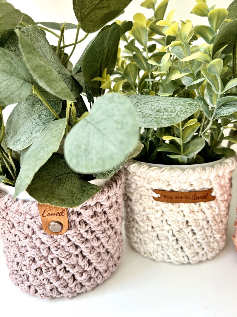May include: Two potted plants with crocheted covers. The plant on the left has a pink cover with a brown leather tag that says "Loved". The plant on the right has a white cover with a brown leather tag that says "You are so loved".