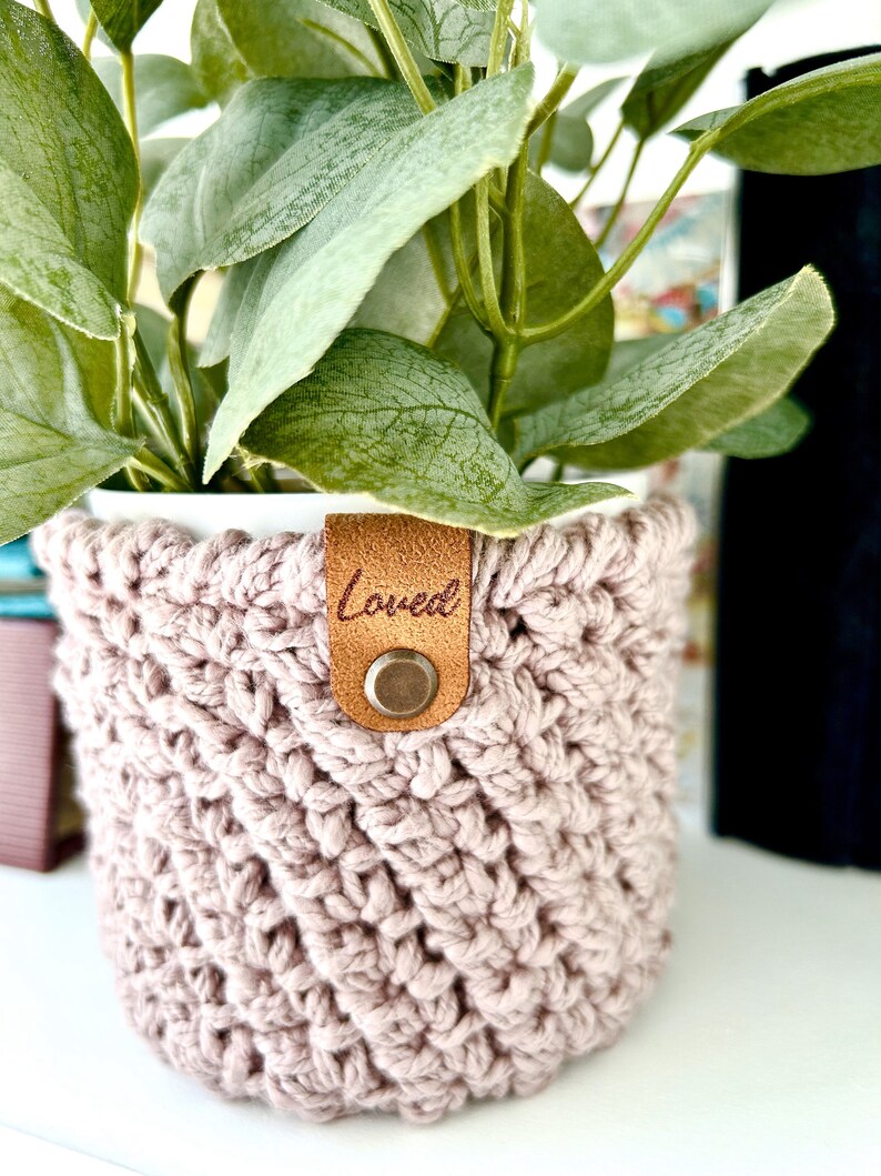 May include: A pink crocheted plant pot with a brown leather tag that says "Loved".