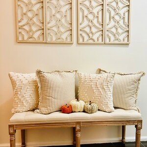 Peut inclure: Un banc blanc avec un coussin beige et quatre coussins. Les coussins ont une bordure en frange et un motif ray&eacute; blanc et beige. Trois petites citrouilles tricot&eacute;es sont sur le banc devant les coussins.
