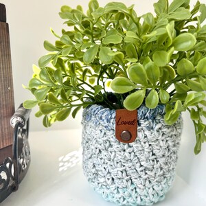 May include: A crocheted plant pot with a blue and green ombre design. The pot has a brown leather tag with the word "Loved" written on it. A small green plant is in the pot.