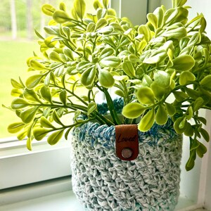 May include: A crocheted plant pot with a blue and white ombre design. The pot has a brown leather tag with the word "Loved" on it. The pot is filled with a green leafy plant.