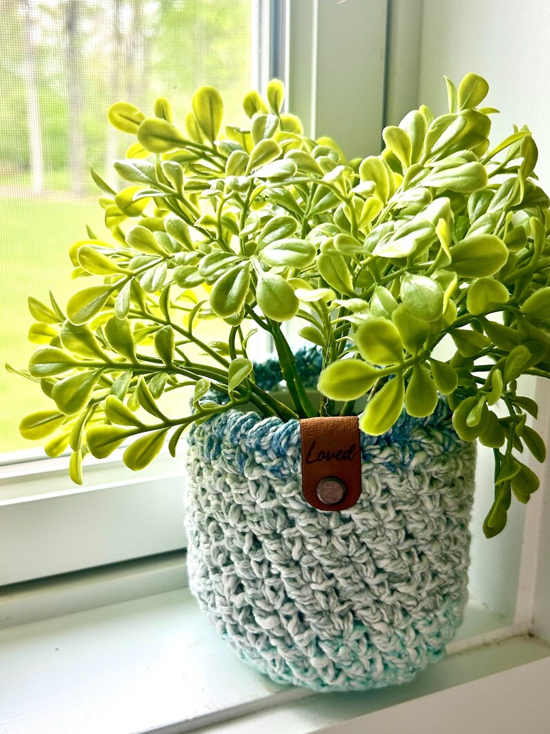 May include: A crocheted plant pot with a blue and white ombre design. The pot has a brown leather tag with the word "Loved" on it. The pot is filled with a green leafy plant.