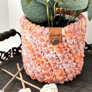 May include: A crocheted plant pot in shades of pink and orange with a brown leather tag that says "Loved".