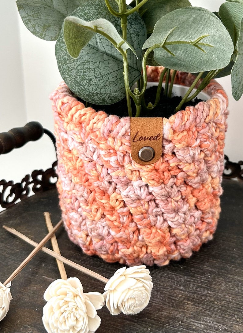 May include: A crocheted plant pot in shades of pink and orange with a brown leather tag that says "Loved".