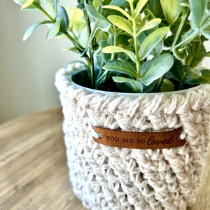May include: A white crocheted plant pot with a brown leather tag that says "you are so loved". The pot is filled with a green leafy plant.