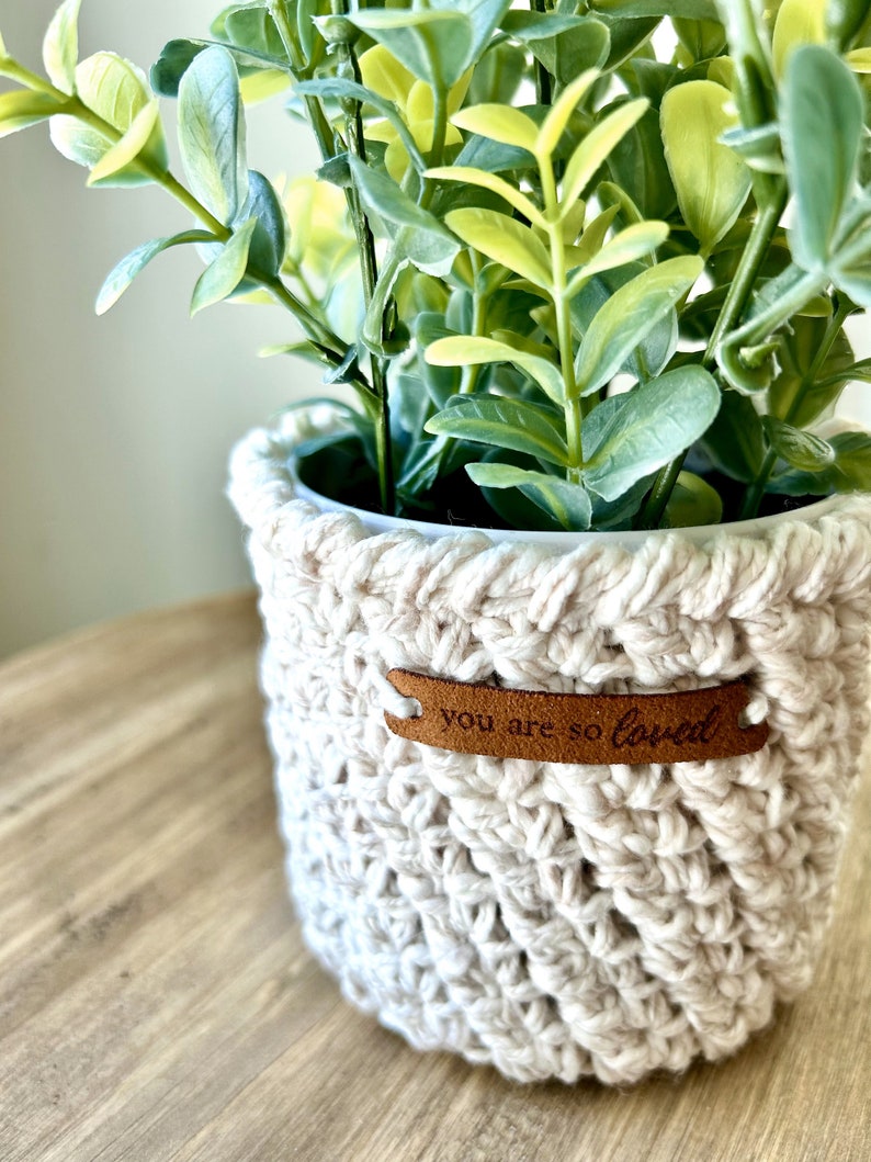 May include: A white crocheted plant pot with a brown leather tag that says "you are so loved". The pot is filled with a green leafy plant.
