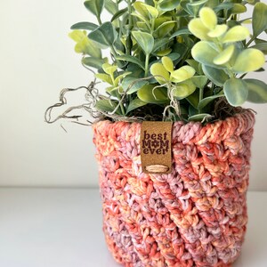 May include: A crocheted plant pot in shades of pink and orange with a brown leather tag that reads "best mom ever".