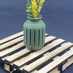 Peut inclure: Un vase en céramique vert avec un motif décoratif posé sur une palette en bois. Le vase est rempli de fleurs jaunes.