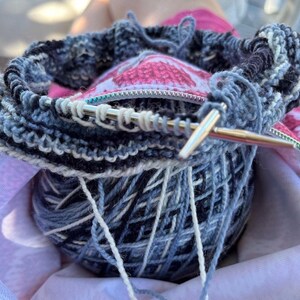 May include: A ball of blue and white yarn is being used to knit a project. The yarn is in a knitting bag with a pink and white floral pattern. A knitting needle is visible in the image.