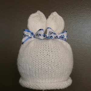 May include: White knitted baby hat with bunny ears and a blue and white ribbon bow.