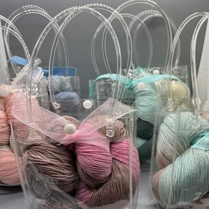 May include: Six clear plastic bags with handles, each containing a skein of yarn. The yarn is in various shades of pink, brown, blue, and white.
