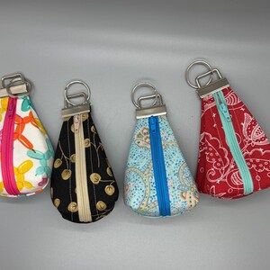 May include: Four teardrop-shaped keychains with metal rings. The keychains feature colorful patterns: a balloon design, black with gold leaves, a blue paisley, and a red bandana print. Each has a zipper closure.
