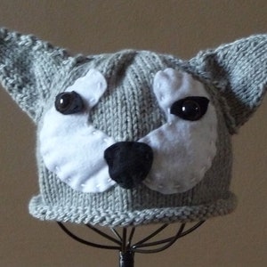 May include: Gray knitted hat with a dog face design. The hat has white felt ears and a black felt nose. The eyes are black buttons.
