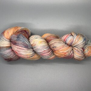 May include: A skein of hand-dyed yarn in a blend of colors including cream, peach, blue, and red. The yarn is tightly wound and appears soft, ideal for knitting or crocheting projects. The color variations create a speckled effect.