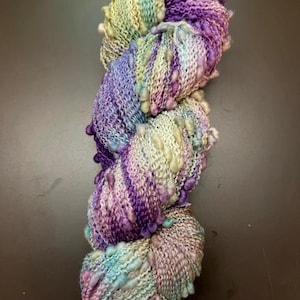 May include: A skein of yarn with a gradient of purple, blue, and white colors. The yarn has small, white pom-poms throughout.