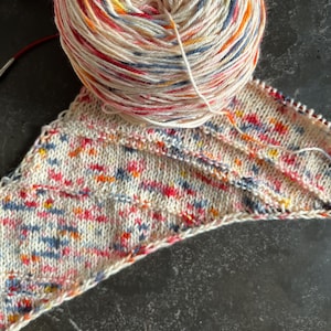 May include: A ball of colorful yarn with a white base and red, orange, yellow, blue, and grey speckles. A knitted piece of fabric in the same colors is partially visible in the background.