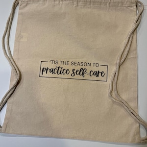 May include: Beige drawstring bag with the text "'TIS THE SEASON TO practice self-care" in a black, rectangular frame. The bag has a simple design and is made of a light-colored fabric. The drawstring is a light beige color.