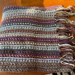 May include: A handmade crochet blanket with a striped pattern in shades of gray, purple, brown, and blue. The blanket has a fringed edge with multiple colors. The blanket is folded and appears soft and warm.