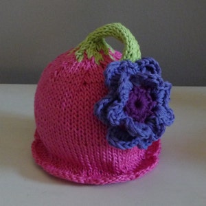 Puede incluir: Un gorro de punto rosa con un tallo verde y una flor morada en la parte superior.