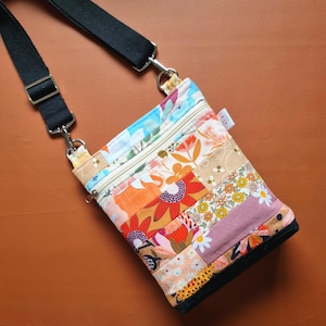 May include: A patchwork crossbody bag with a black adjustable strap. The bag features floral fabric patterns in orange, pink, blue, and beige. It has a black base and a zip closure.