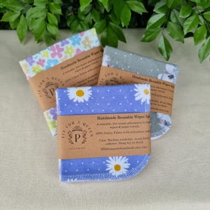 May include: Three stacks of handmade reusable wipes with floral and polka dot patterns. Each stack is wrapped with a brown paper band that includes product information and the brand logo. The wipes are a low-waste alternative to paper towels.