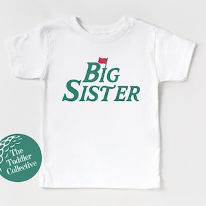 May include: White toddler t-shirt with the words "Big Sister" in green script, topped with a red flag graphic. A green and white logo for "The Toddler Collective" is on the lower left.