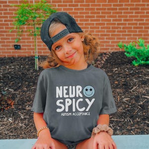 May include: A child wearing a grey t-shirt with the text "Neuro Spicy Autism Acceptance" and a black baseball cap. The child is smiling and looking at the camera.