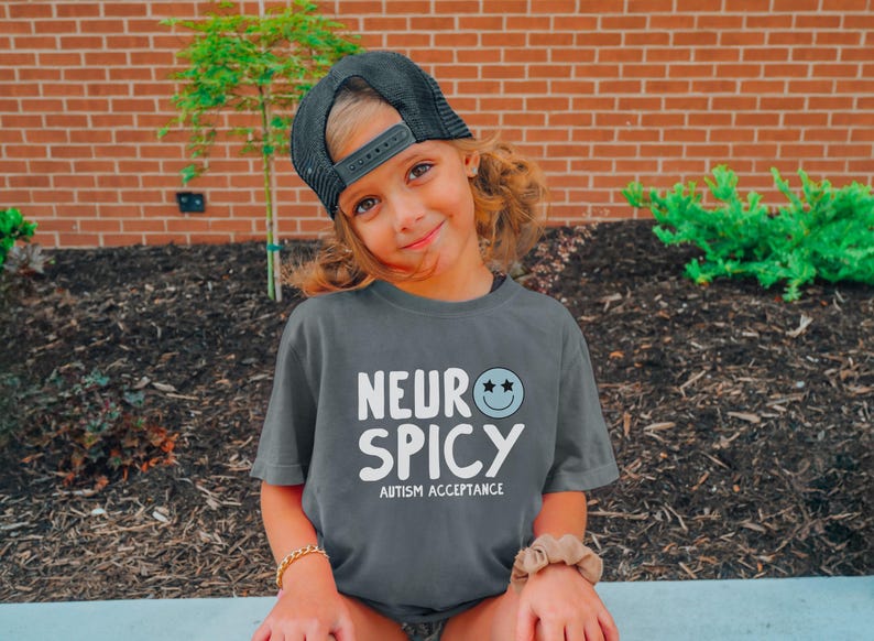 May include: A child wearing a grey t-shirt with the text "Neuro Spicy Autism Acceptance" and a black baseball cap. The child is smiling and looking at the camera.