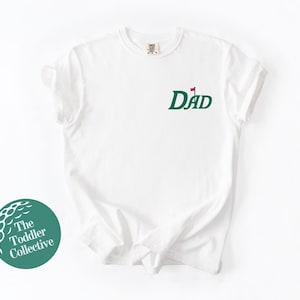 May include: White t-shirt with a green golf-themed "DAD" graphic. The design includes a golf flag. The shirt has a crew neck and short sleeves. The Toddler Collective logo is visible in the lower left corner.
