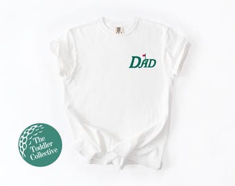 May include: White t-shirt featuring a green golf-themed "DAD" graphic with a golf flag. The shirt has a crew neck and short sleeves. The Toddler Collective logo is visible in the lower left corner. The shirt is made of cotton.