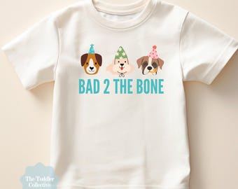 May include: White toddler t-shirt featuring three cartoon dog faces wearing party hats. The text "BAD 2 THE BONE" is printed in teal below the dogs. The shirt is from "The Toddler Collective".