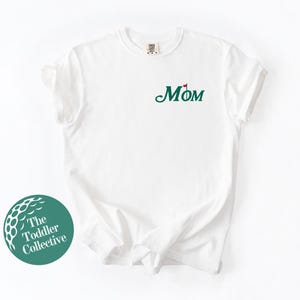 May include: White t-shirt with a green "MOM" graphic with a red golf flag on the "O".